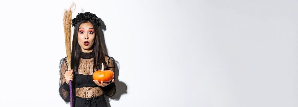Image of surprised asian girl gasping wondered and stare at camera, wearing witch costume on halloween, holding broom and pumpkin, white background.