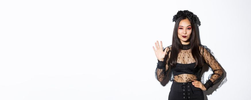 Image of confident beautiful asian woman in halloween costume showing five fingers, raising hand to say hello, standing over white background.