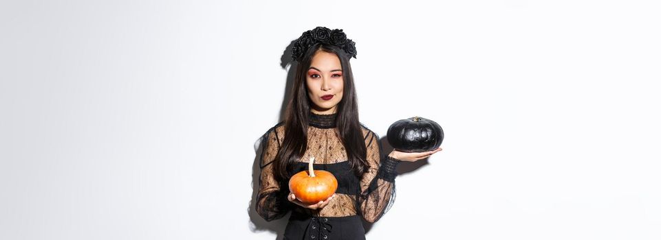 Sassy attractive asian girl in witch costume, holding pumpkins and looking at camera, standing over white background.