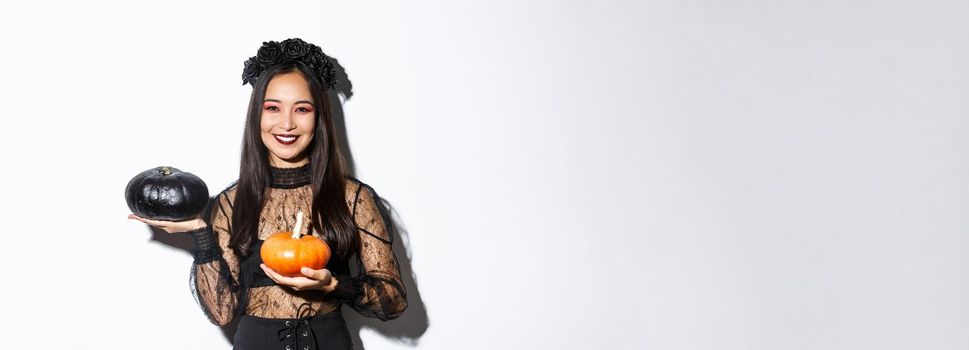 Image of attractive korean girl celebrating halloween in gothic lace dress, impersonating witch and holding two pumpkins.
