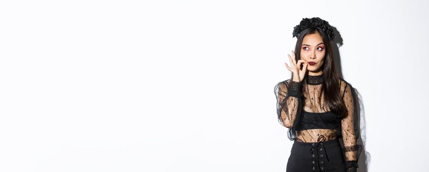 Young gothic woman in black lace dress, dressed-up like witch for halloween party, making seal on lips, zipping mouth as making promise keep secret, standing over white background.