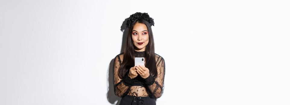 Thoughtful pretty asian girl in gothic elegant dress holding smartphone while thinking, looking at upper left corner, standing over white background.