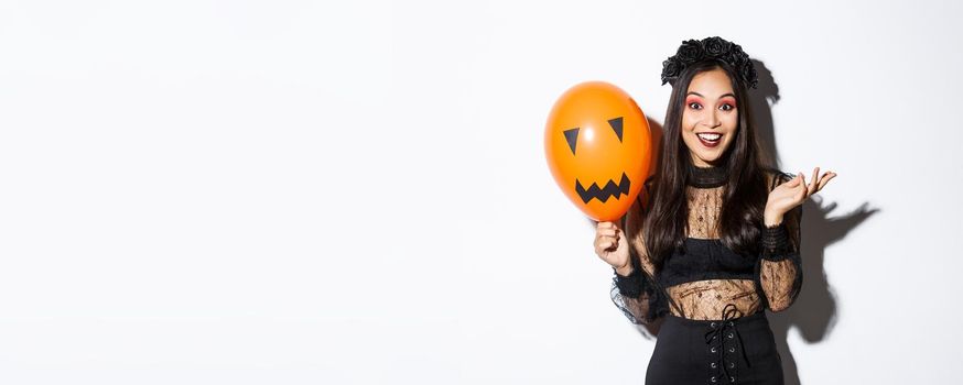 Image of beautiful asian woman celebrating halloween, wearing witch costume and gothic makeup, talking with orange balloon.