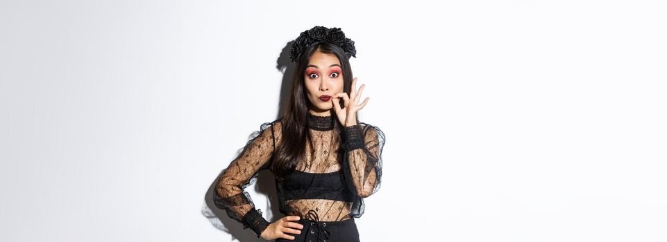 Pretty asian girl in witch costume making promise, seal lips and keep secret, zip mouth, censored information, standing over white background in halloween outfit.