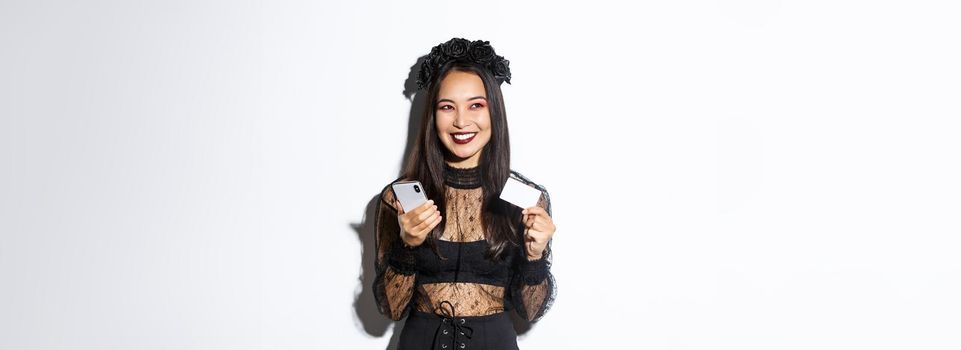 Image of beautiful asian woman in gothic lace dress and black wreath, looking aside pleased and smiling, holding mobile phone with credit card.