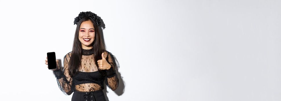 Image of happy and satisfied asian woman in halloween costume showing thumbs-up and demonstrating mobile phone screen, smiling pleased, standing over white background.