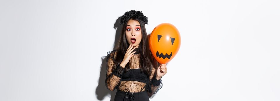 Portrait of surprised asian woman in halloween costume, dressed-up as witch, holding orange balloon with scary face.