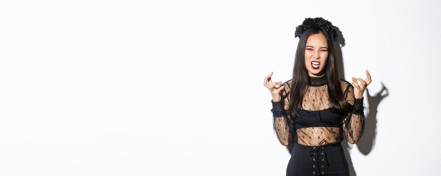 Image of woman in halloween costume looking like angry evil witch, cursing and clenching fists mad. Female in black dress and wreath trick or treating, standing white background.