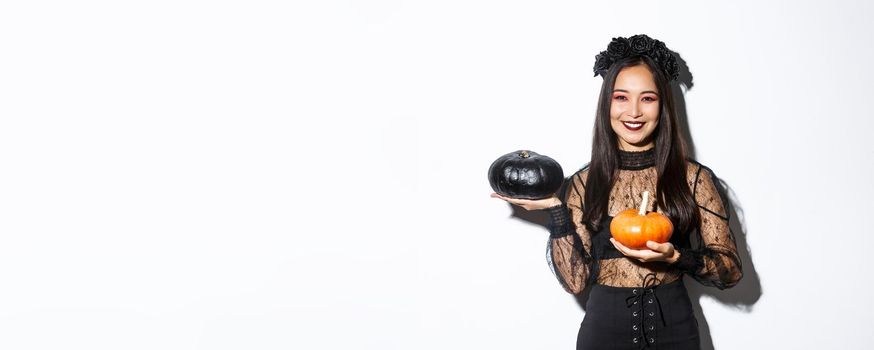 Image of attractive korean girl celebrating halloween in gothic lace dress, impersonating witch and holding two pumpkins.