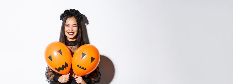 Image of asian girl in evil witch costume holding two orange balloons with scary faces, celebrating halloween, standing over white background.