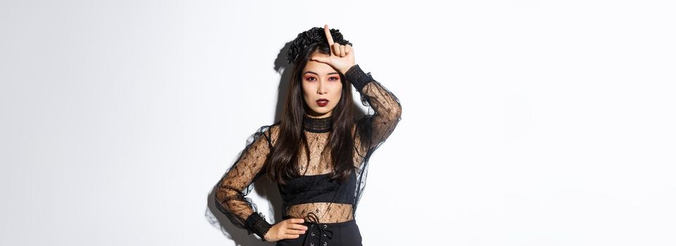 Image of arrogant sexy asian woman in witch costume showing loser gesture on forehead, mocking someone at halloween party, standing over white background.
