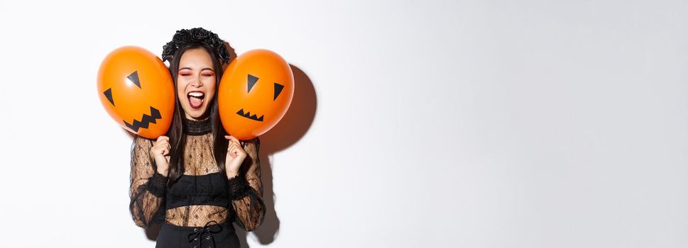 Image of asian girl in evil witch costume holding two orange balloons with scary faces, celebrating halloween, standing over white background.