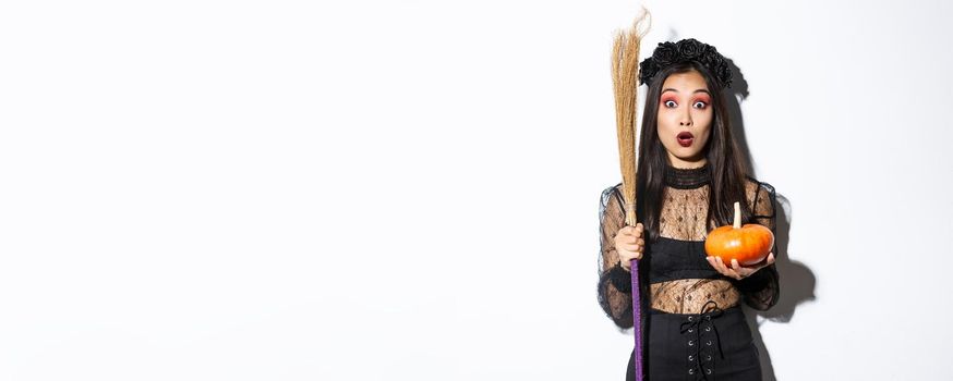 Image of surprised asian girl gasping wondered and stare at camera, wearing witch costume on halloween, holding broom and pumpkin, white background.