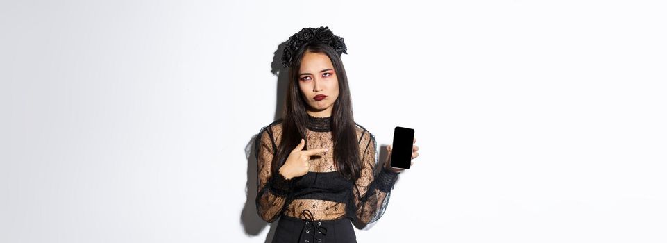Image of annoyed and reluctant asian girl in witch costume, roll eyes and pouting while pointing finger at mobile phone screen, standing over white background.
