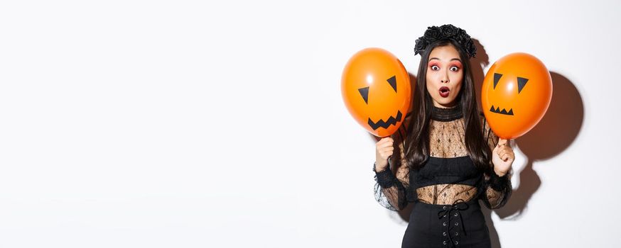 Image of surprised asian woman in witch costume celebrating halloween, holding balloons with scary faces, standing over white background.