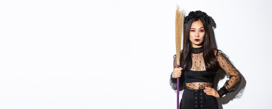 Beautiful asian woman in gothic lace dress and black wreath, holding broom and looking suspicious at camera, standing over white background.