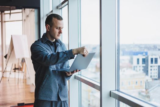 Portrait of a smart intelligent male manager, he is holding a laptop for research in the office, a business man in elegant clothes playing on the touchpad. Standing in the office by a large window