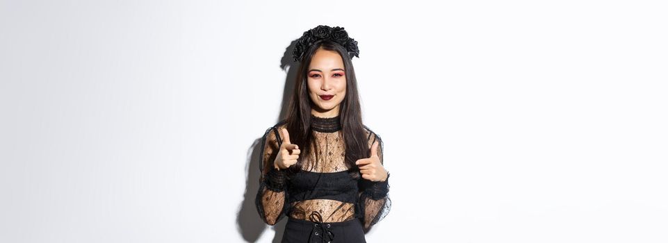 Cheeky and happy attractive woman in witch outfit pointing fingers at camera and smiling, making compliment or praising something good, standing in halloween costume over white background.
