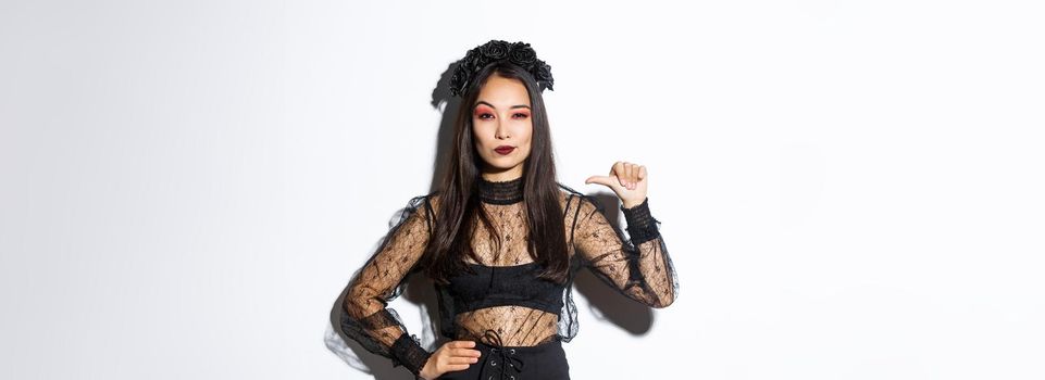 Confident stylish asian woman looking determined, wearing black lace dress for halloween party, pointing at herself sassy, standing over white background.