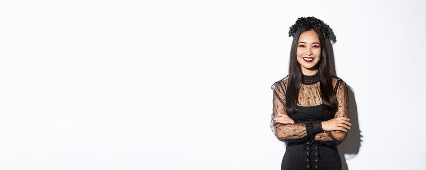 Smiling beautiful asian woman celebrating halloween, wearing black wreath and dress, gothic makeup, looking happy at camera. Female celebrating autumn event, trick or treating in costume.