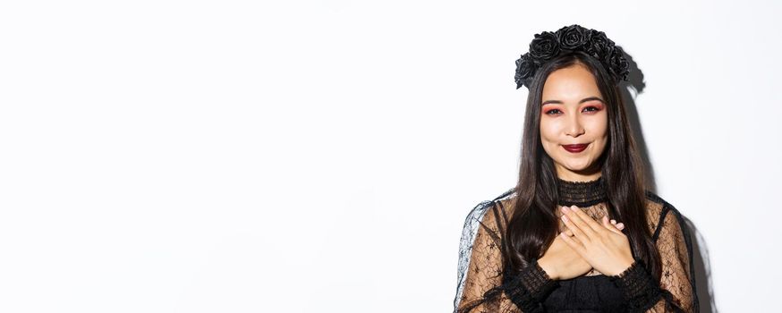 Close-up of thankful smiling asian woman looking grateful with hands over chest, standing in witch costume over white background.