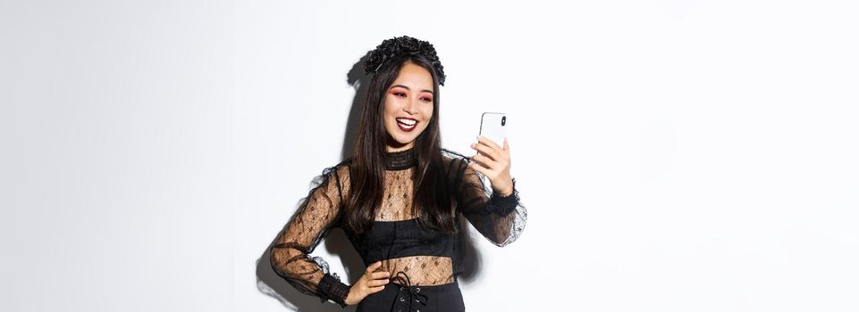 Portrait of happy beautiful asian woman in halloween costume smiling and looking at mobile phone screen, having video call, standing over white background.