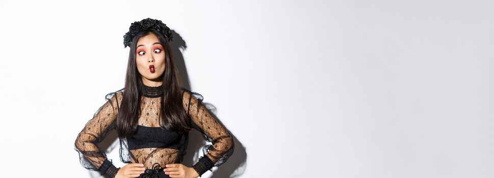 Image of funny asian girl celebrating halloween, wearing gothic lace dress to impersonate witch, showing silly faces and having fun, standing over white background.