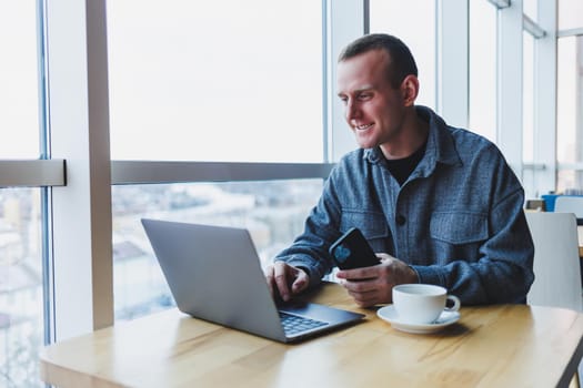 Successful happy business man is sitting at a table in a cafe, holding a cup of coffee and using a laptop.