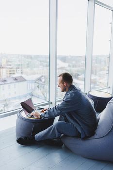 Break. Smiling young man relaxing on bean bag chair while sitting at table and relaxing using laptop, happy businessman leaning back, enjoying work, feeling satisfied