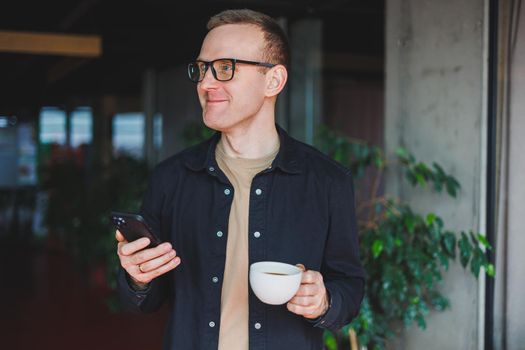 Portrait of a happy successful man in optical glasses holding a modern mobile phone and coffee in his hands, a cheerful blogger with a digital smartphone posing in a cafe