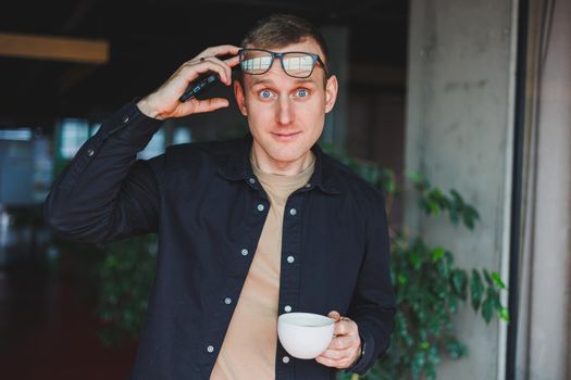 Portrait of a happy successful man in optical glasses holding a modern mobile phone and coffee in his hands, a cheerful blogger with a digital smartphone posing in a cafe