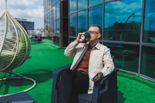 Confident young businessman, dressed in formal wear and stylish glasses, calls on a smartphone while sitting with coffee, on the terrace of a modern office building with a glass wall