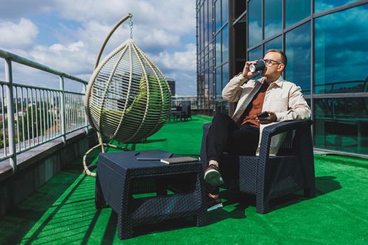 Confident young businessman, dressed in formal wear and stylish glasses, calls on a smartphone while sitting with coffee, on the terrace of a modern office building with a glass wall