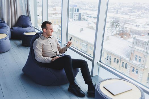 Successful male freelancer sitting in modern workspace and checking notifications on mobile phone in casual clothes. Remote work.