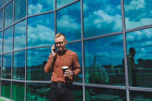 Attractive fashionable male businessman in a stylish shirt and trousers talking on the phone near the office building and drinking coffee, modern glass office