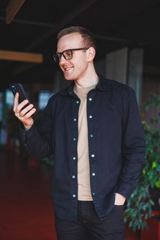 Half-length portrait of happy man in optical glasses holding modern mobile phone and smiling at camera, cheerful blogger with digital smartphone posing in coworking space