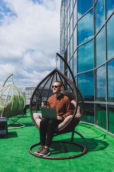 A businessman in formal wear sits on an armchair and looks at a laptop connected to a 4g wireless network on the terrace of a modern office while working remotely