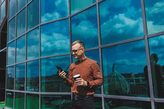 Attractive fashionable male businessman in a stylish shirt and trousers talking on the phone near the office building and drinking coffee, modern glass office