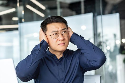 Noise in the office, Asian worker businessman covers his ears with hands from noise, man works inside office building complains about loud sound.