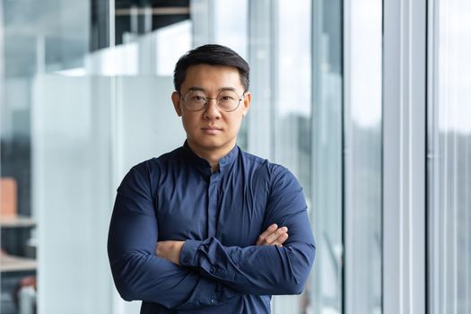 Portrait of successful and serious asian boss in shirt, man looking at camera with crossed arms inside modern office building near window.