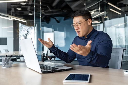 Frustrated and angry asian boss yelling at laptop, failure man working inside twisted office building at desk.