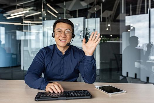 Webcam view, portrait of successful tech support and customer service worker, man smiling and looking at camera waving greeting hello, asian working inside modern office building.