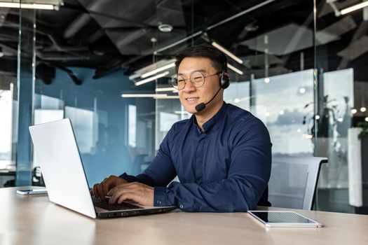 Happy and smiling worker with headset for video call, Asian tech support man talking to online service users, businessman using laptop for online customer support.