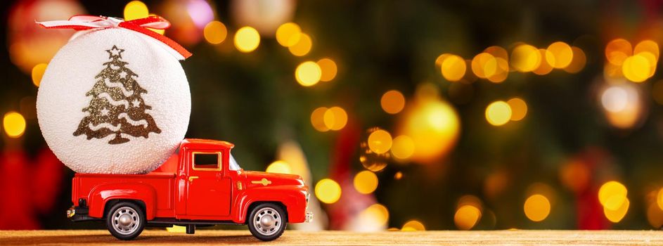 Banner photo of red small retro toy truck with white christmas ball on background of Christmas tree lights. Christmas, New Year concept. Copy space.