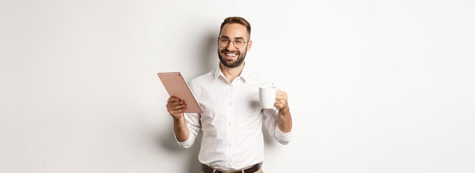 Satisfied boss drinking tea and using digital tablet, reading or working, standing over white background.