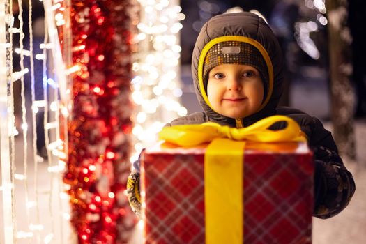 Happy child in a red Santa Claus hat with wrapped Christmas gift near shining light bulbs in winter evening. The concept of Christmas and New Year.