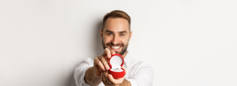 Close-up of handsome man asking to marry him, focus on box with wedding ring, concept of proposal and relationship, white background.