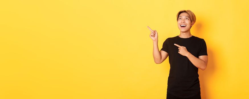 Portrait of pleased handsome asian guy in black t-shirt, pointing fingers and looking upper left corner with satisfied smile, yellow background.