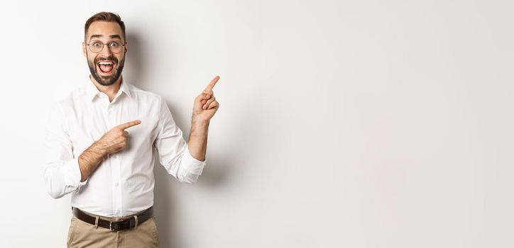 Excited handsome man pointing fingers at upper right corner, showing logo, standing over white background.
