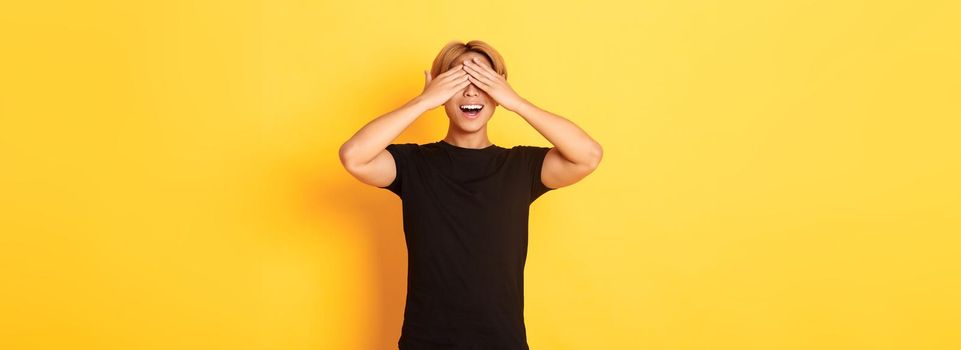 Portrait of amused and excited smiling blond guy shut eyes with palms, waiting for surprise, standing yellow background.